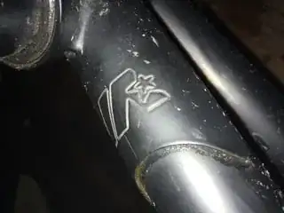 Close-up of the logo on the lug at the front of the top tube. The logo is a stylized "M" formed of three strokes, with a five- pointed star partially inset in the trailing stroke.
